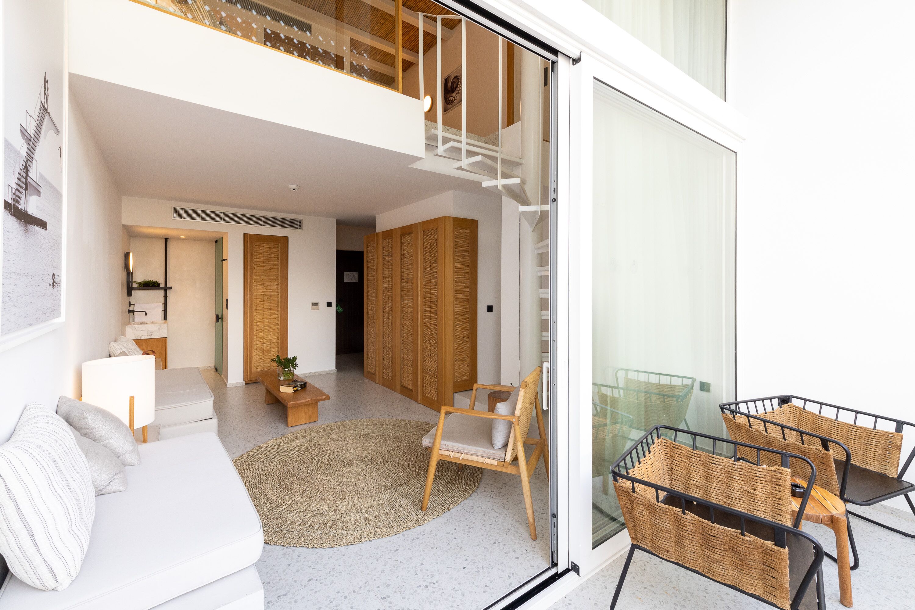 Una sala de estar moderna con un sofá blanco, una silla de madera y una mesa de vidrio. Se ve una escalera que sube a un nivel superior.