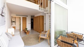 Una sala de estar moderna con un sofá blanco, una silla de madera y una mesa de vidrio. Se ve una escalera que sube a un nivel superior.
