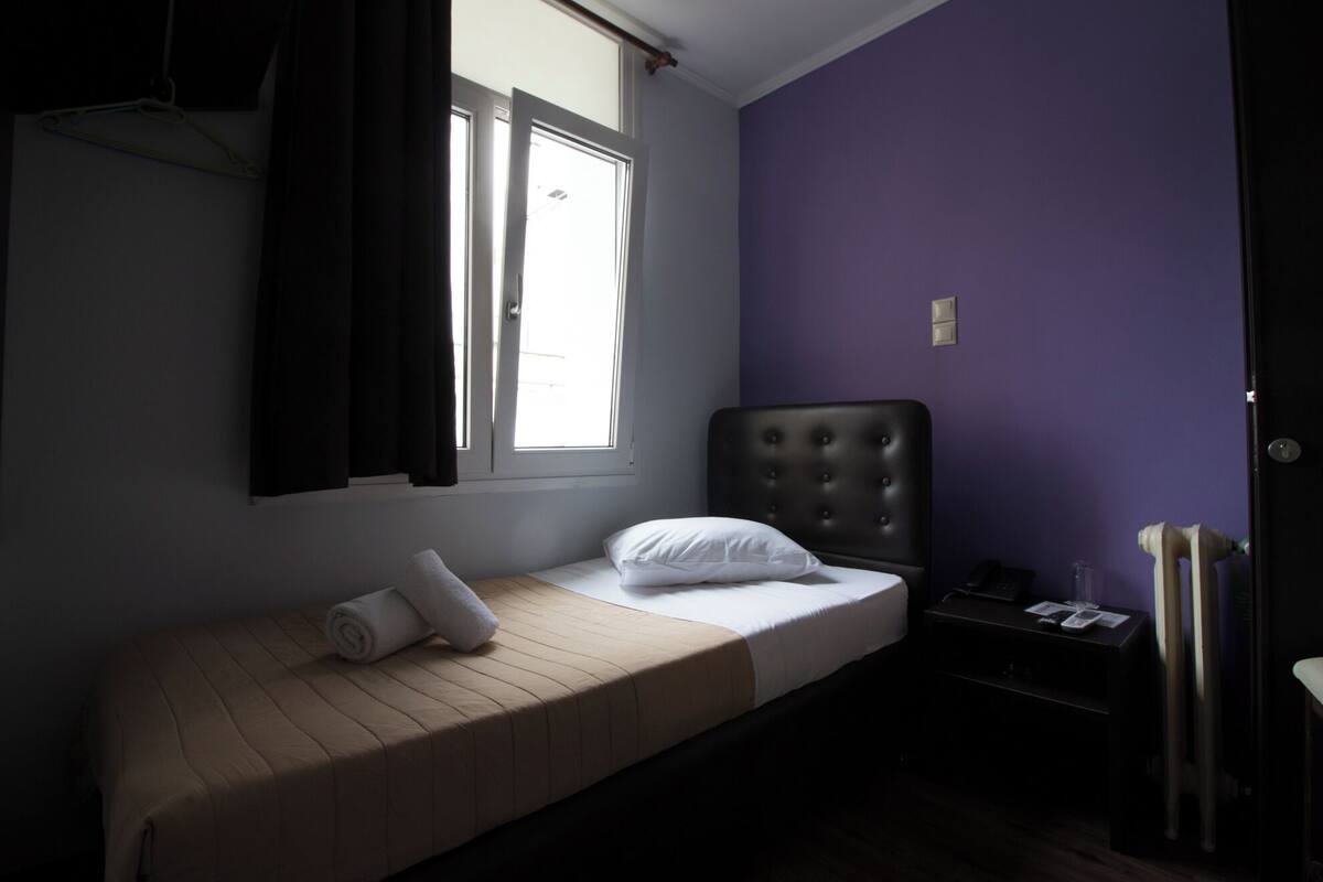 Una habitación pequeña con cama individual, ventana, mesita de noche y una silla.