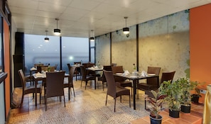 A restaurant with tables and chairs, large windows, and potted plants. - The Pamposh New Delhi (New Delhi)