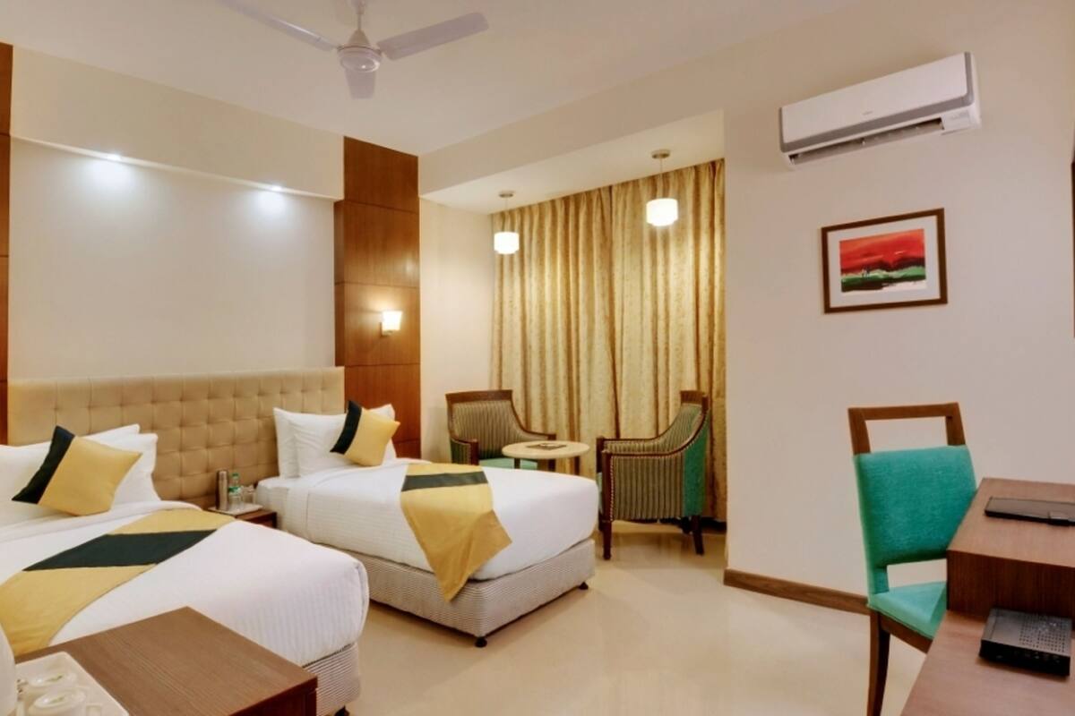A hotel room with two beds, a desk, a chair, a table, a ceiling fan, an air conditioner, and a painting on the wall.