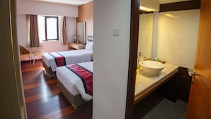 Ein Hotelzimmer mit zwei Betten, einem Schreibtisch, einem Stuhl, einem fenster mit Vorhängen und einem Badezimmer mit Waschbecken und Spiegel.