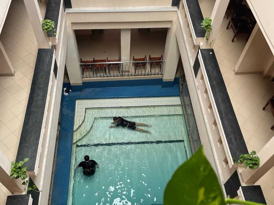 Uma piscina interior com duas pessoas nadando, cercada por um edifício com vários níveis, varandas e plantas em vasos.