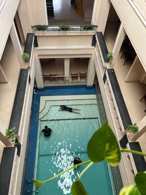 An indoor swimming pool with two people swimming, surrounded by a multi-level building with balconies and potted plants. - Malioboro Inn Yogyakarta (Yogyakarta)