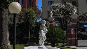 Una fuente con una estatua en un jardín ajardinado, rodeado de árboles y un edificio al fondo.