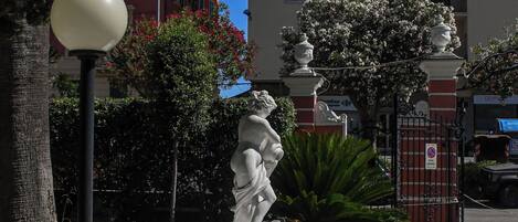 A fountain with a statue in a landscaped garden area, surrounded by trees and a building in the background.