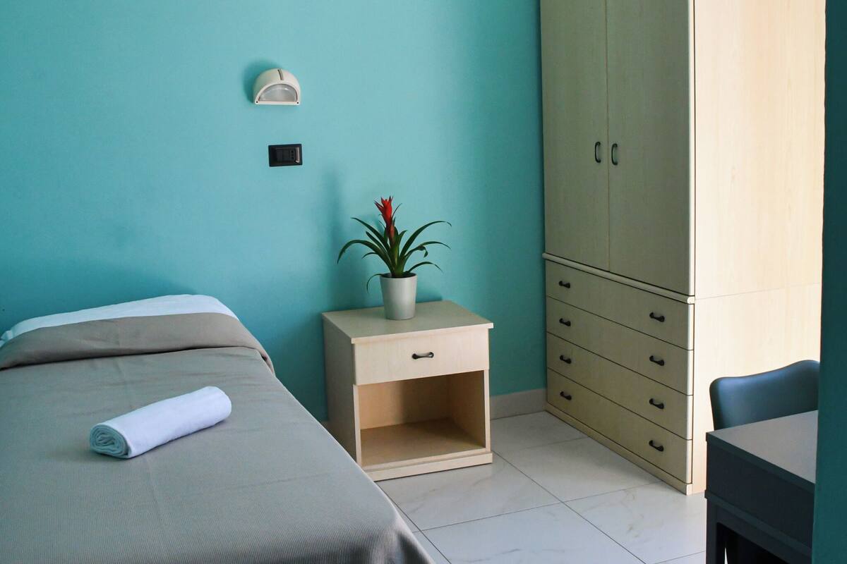 Une pièce aux murs de couleur turquoise, un lit recouvert d’une couverture beige, une armoire en bois, une petite table de chevet avec une plante et une chaise.
