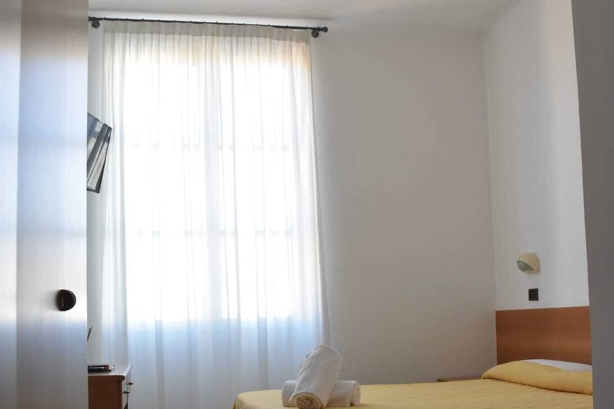 Une chambre d’hôtel avec un lit, une commode en bois, une fenêtre avec des rideaux légers et un mur sobre.