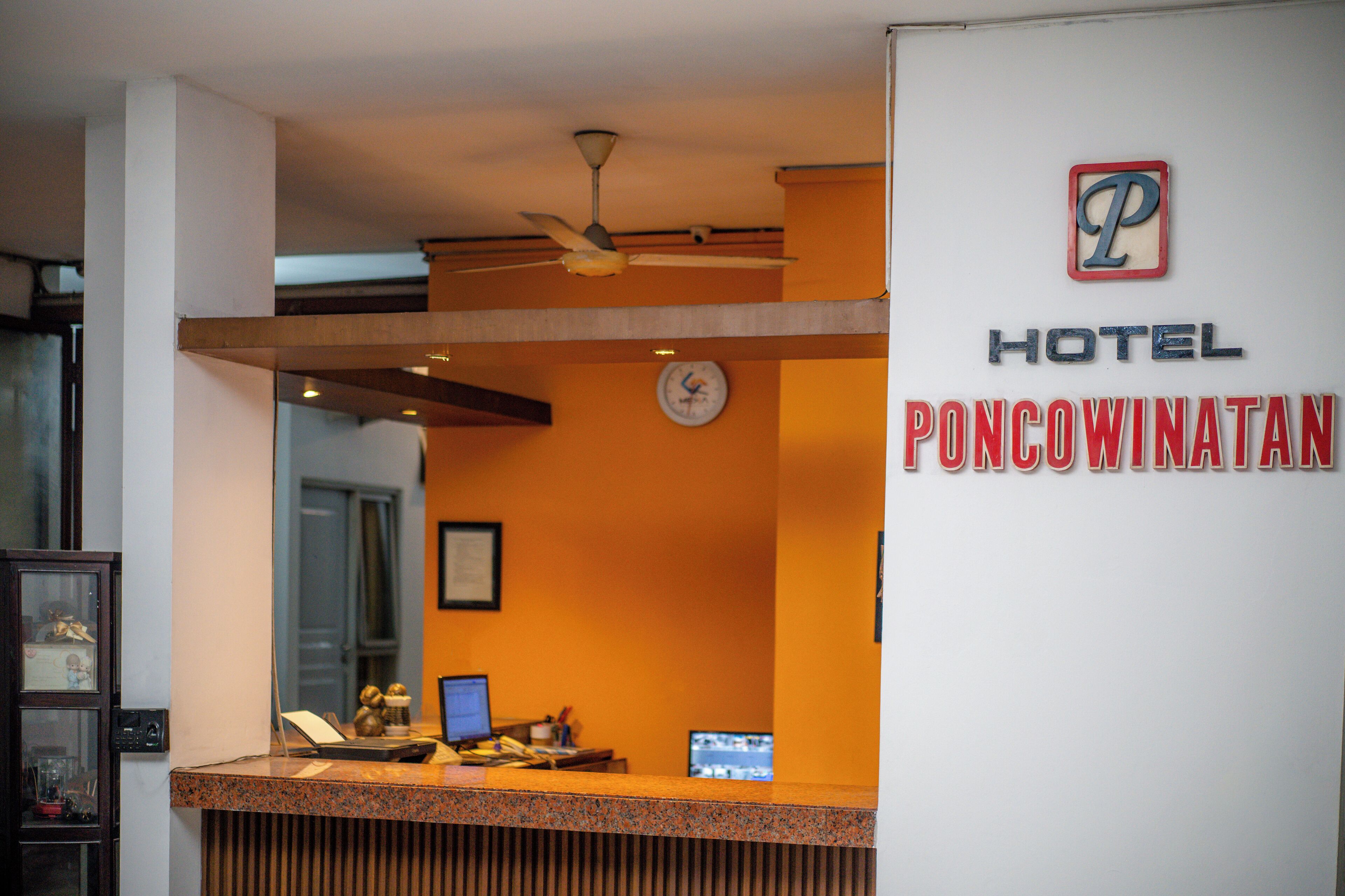 Un comptoir d’accueil avec une horloge, un ventilateur de plafond et une enseigne indiquant l’hôtel Poncowinatam.