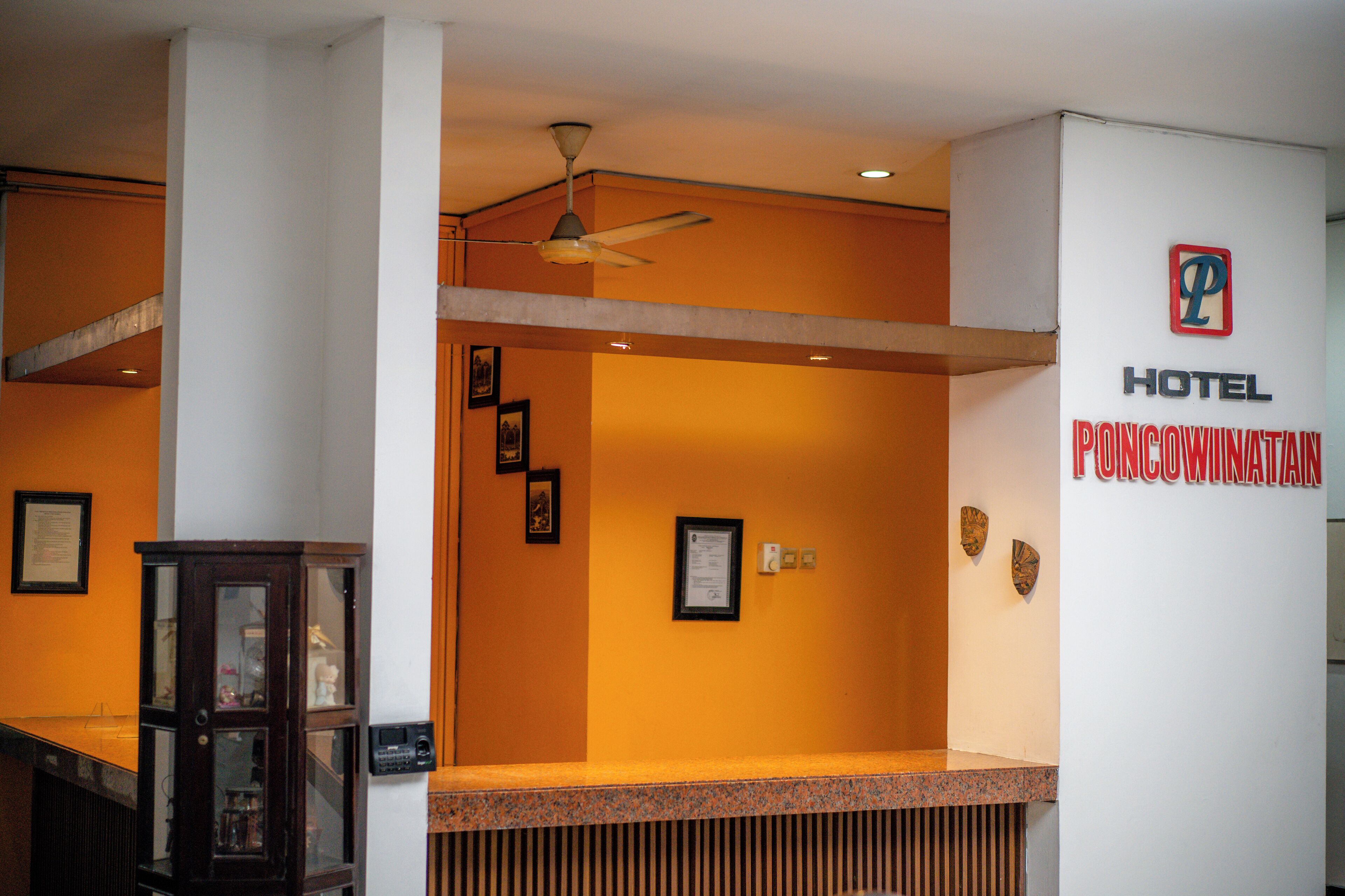Ein Empfangsbereich mit Theke, Deckenventilator, gerahmten Bildern an der Wand und einem Schild für "Hotel Poncowinatan".
