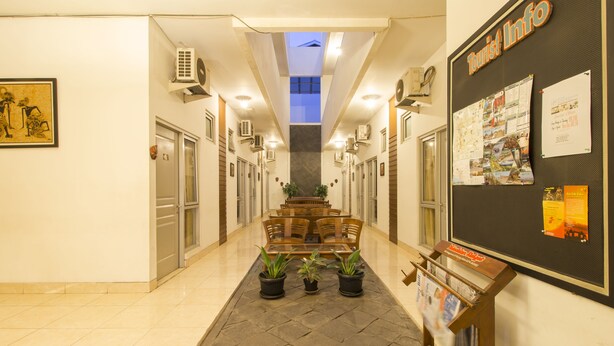Un pasillo de hotel con un área de estar central, unidades de aire acondicionado, un tablón de anuncios con información turística y una obra de arte enmarcada en la pared.