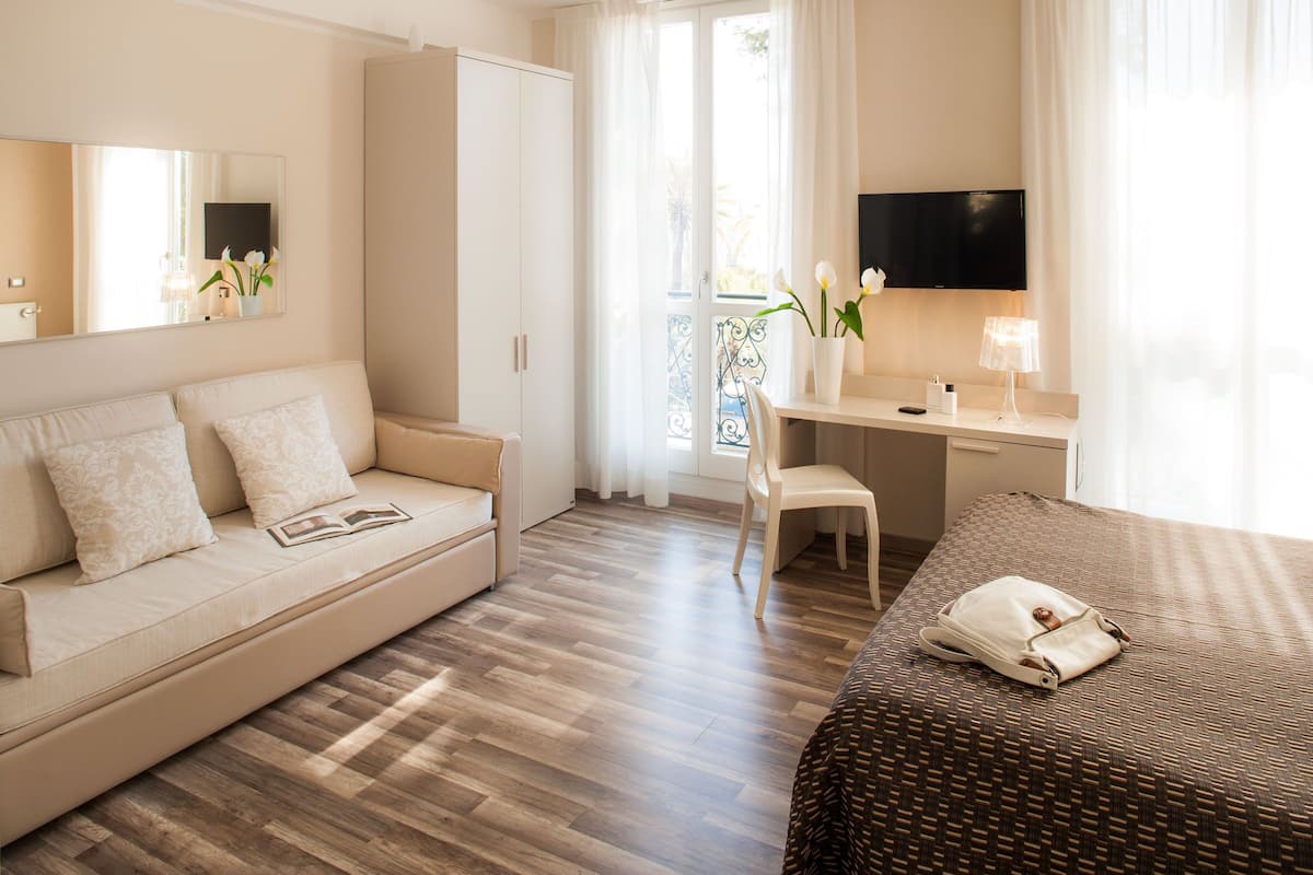 A hotel room with a sofa, a desk, a chair, a bed, a TV, and a window with curtains.