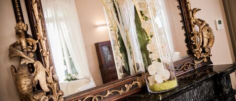 Un espejo con detalles ornamentales dorados, un florero con flores blancas y una chimenea de mármol.
