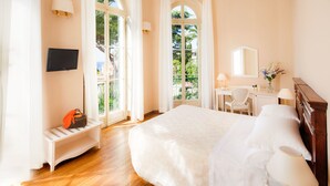 A hotel room with a large bed, a desk with a chair, a TV mounted on the wall, and a window with curtains. - Villa Sylva & Spa (Sanremo)