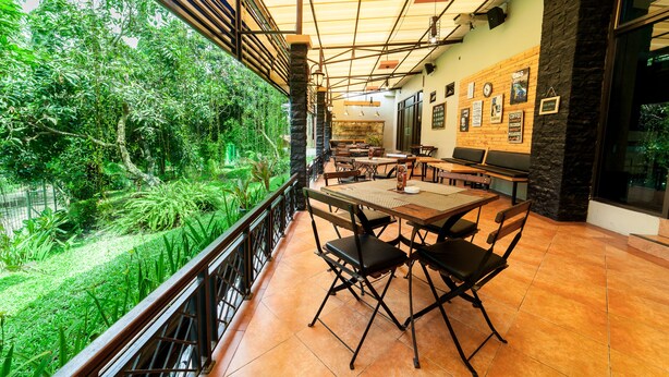 A terrace with a view of lush greenery, wooden tables and chairs, and a stone wall with framed pictures. - MMUGM Hotel Yogyakarta (Depok)