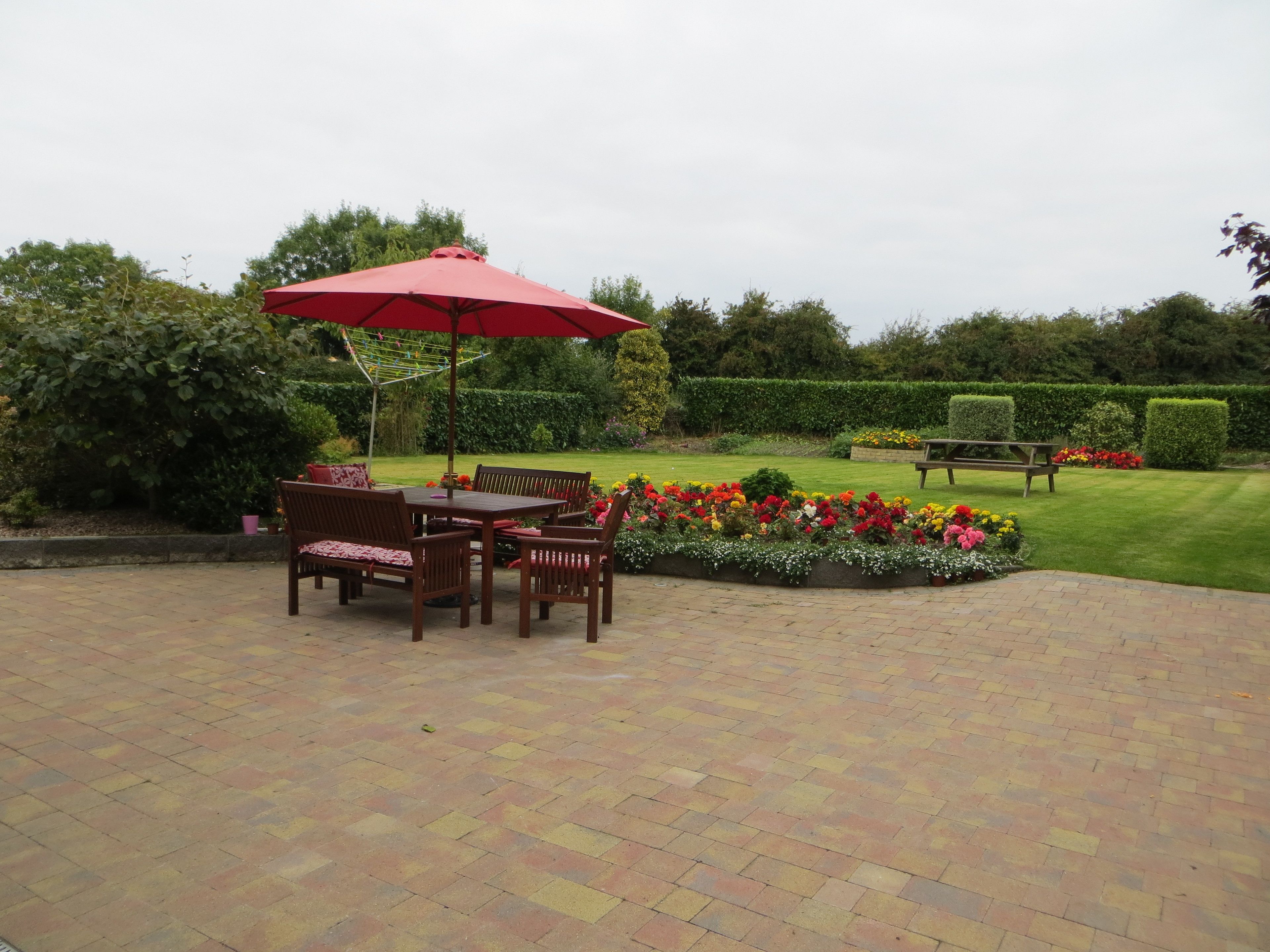 A patio area with a table and chairs under a red umbrella, surrounded by flower beds and a neatly trimmed hedge.