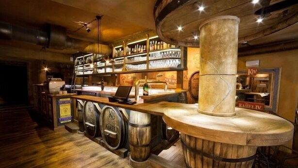 Uma área de bar com barris de madeira, prateleiras com garrafas e um balcão com um laptop.