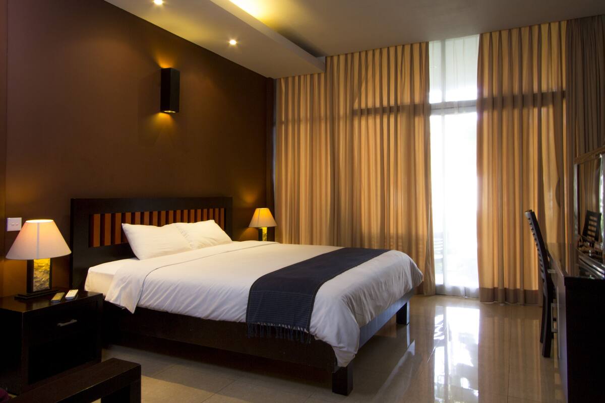 A hotel room with a large bed, two bedside lamps, a piano, and a view of the outdoors through curtains.