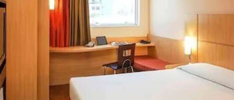 A hotel room with a bed, a desk with a chair, a laptop, and a window with curtains.