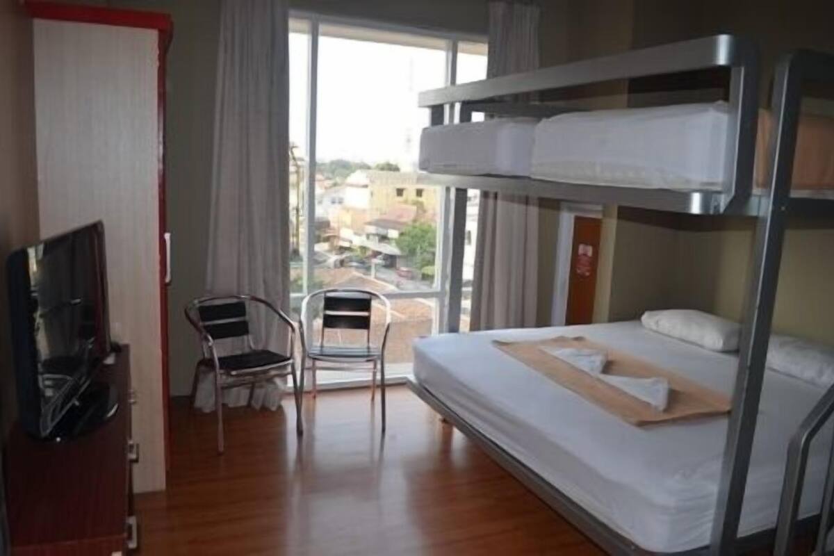 A hotel room with a bunk bed, a desk with a chair, a television, and a window with a view of buildings.