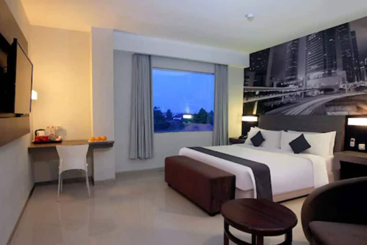 Hotelkamer met een groot bed, een bureau met stoel, een bijzettafeltje en uitzicht op een stadsbeeld in de schemering.