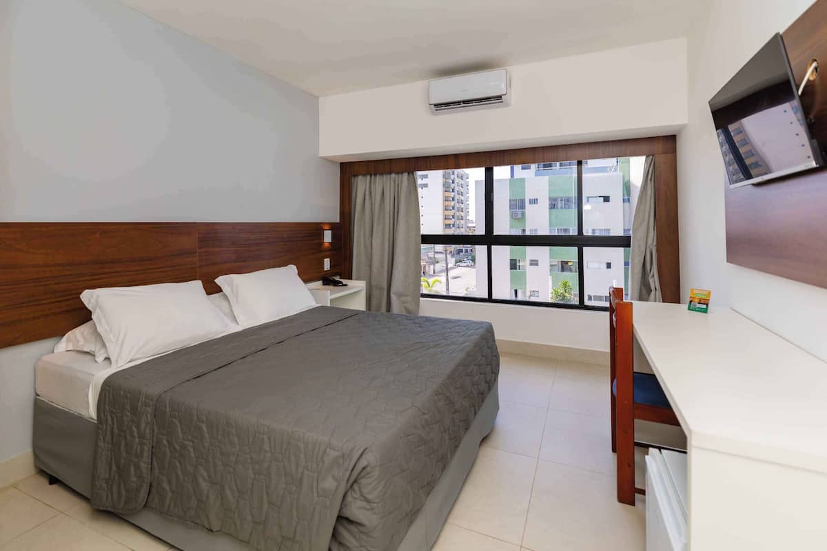 Een hotelkamer met een bed, een bureau, een stoel, een raam met gordijnen en een airconditioning.