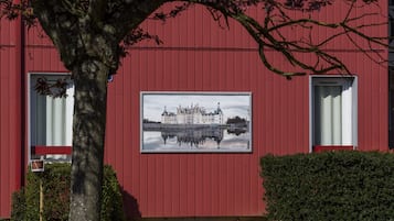 A red building with a framed picture on the wall, a tree in front, and a hedge to the right.