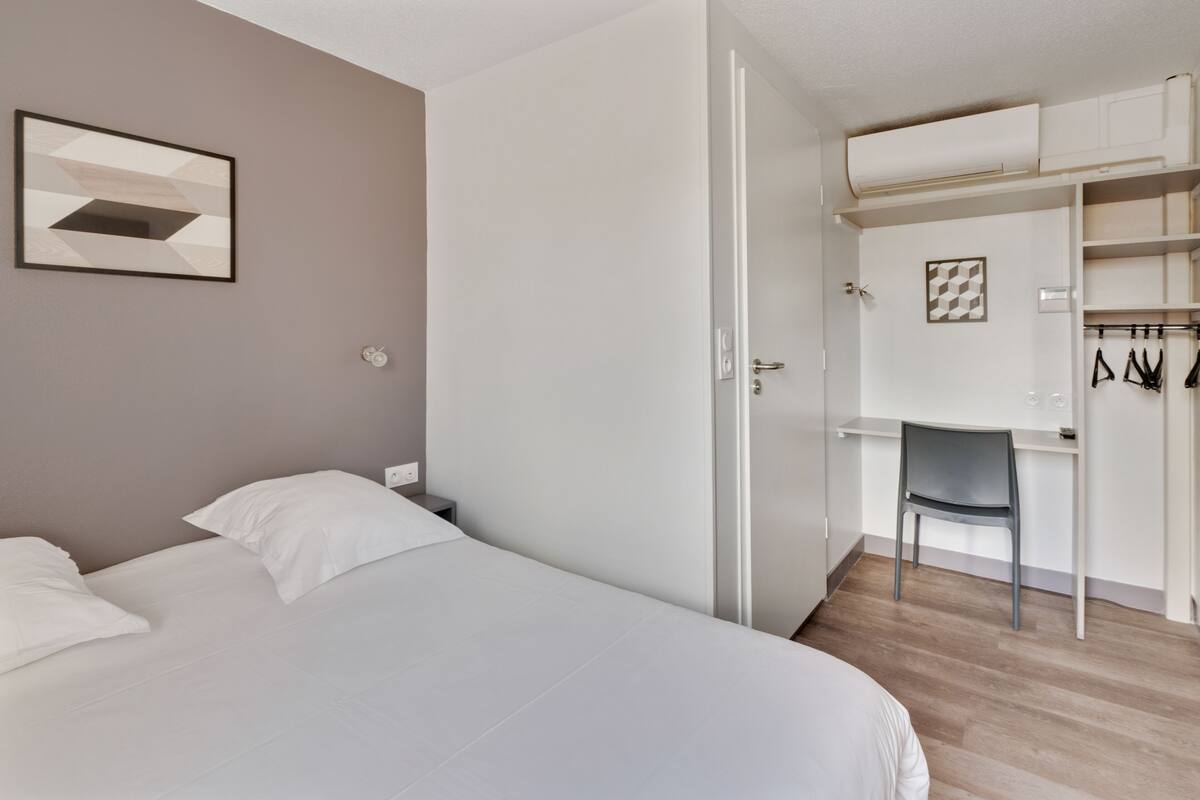 Une chambre à coucher avec un lit, un bureau et une chaise. On y trouve également une armoire où sont suspendus des vêtements.