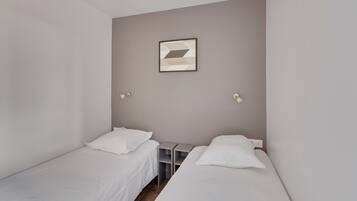 Une petite chambre propre, avec deux lits simples, une petite table et un luminaire fixé au mur.