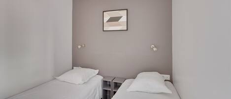 A small, clean room with two single beds, a small table, and a wall-mounted light fixture.