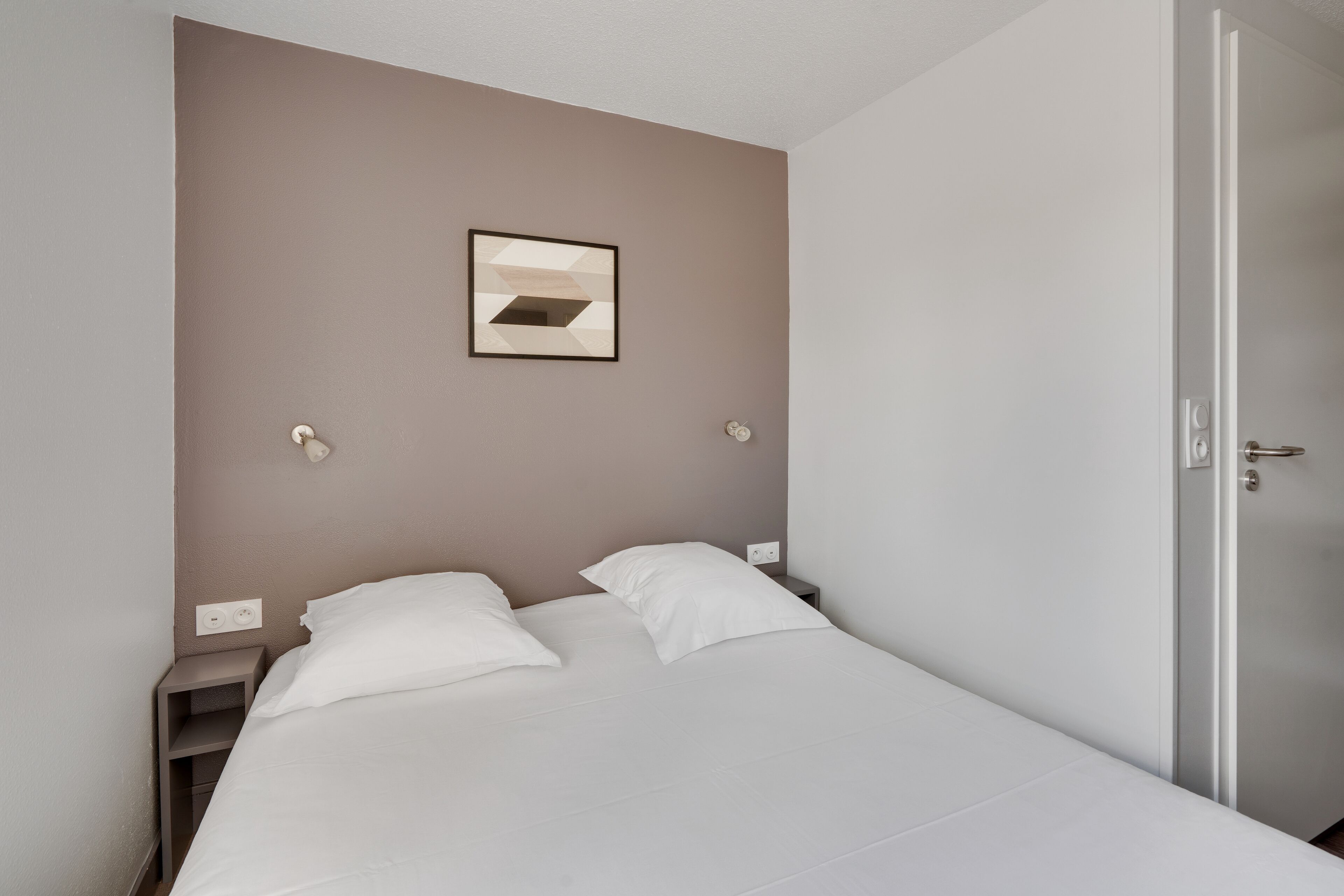 Un lit bien fait, avec du linge de lit blanc, dans une pièce dont un mur est peint dans une teinte marron, avec une petite table de chevet et une porte sur le côté droit.