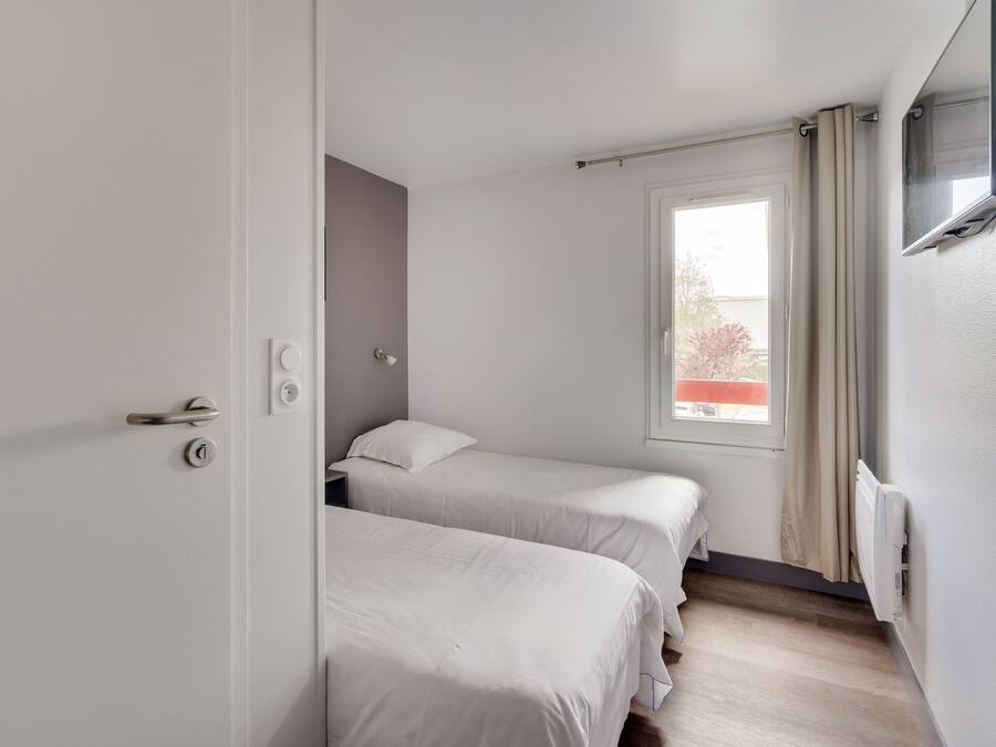 A small, clean room with two single beds, a window with curtains, a TV mounted on the wall, and a door with a handle.