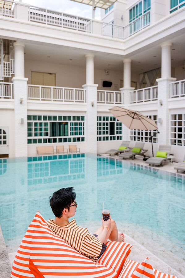A person lounging by a pool with a drink, surrounded by a building with multiple balconies and windows.
