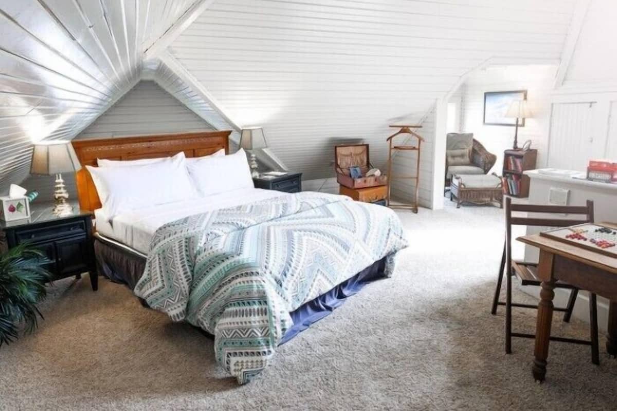 A bedroom with a sloped ceiling, a bed with patterned bedding, a nightstand, a chair, a table, and a lamp.