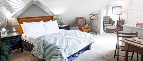 Un dormitorio con techo inclinado, una cama con ropa de cama estampada, una mesita de noche, una silla, una mesa y una lámpara.