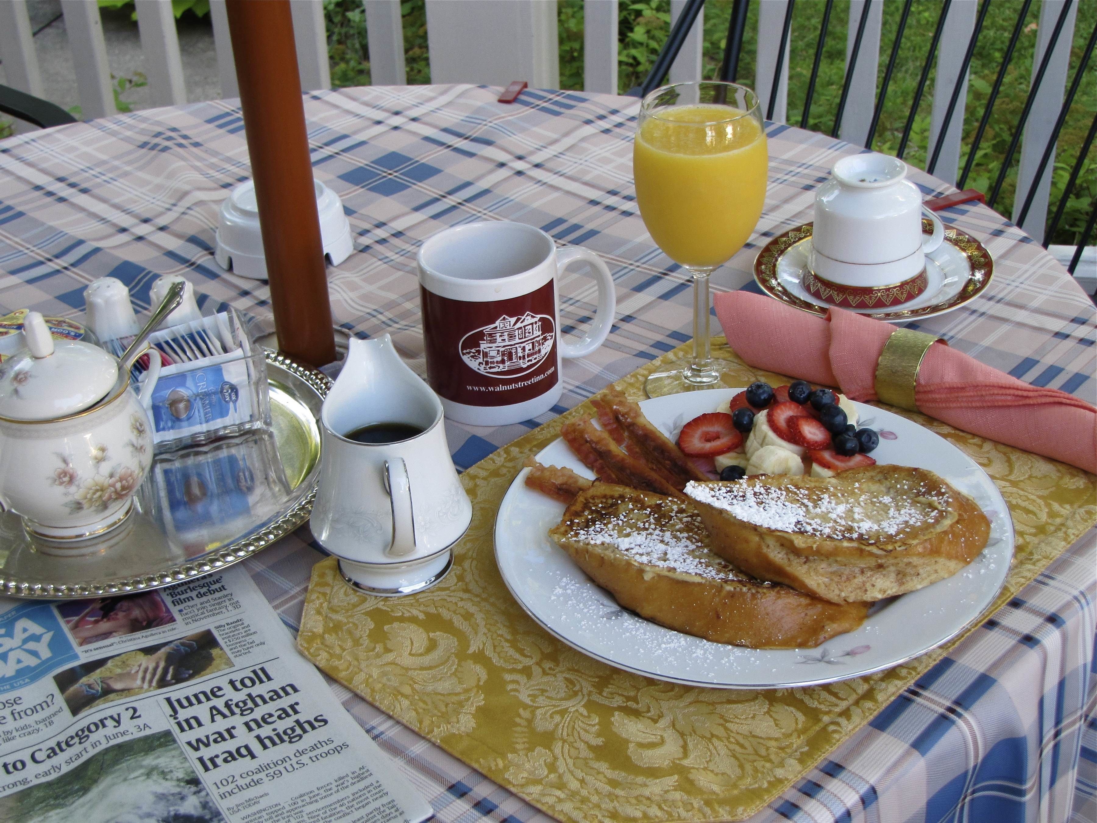 A breakfast table with a newspaper, a glass of orange juice, a cup of coffee, a teapot, and a plate of French toast with berries and powdered sugar.