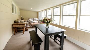 A spacious room with a large window, a dark wooden table, a bench, and a wicker sofa.