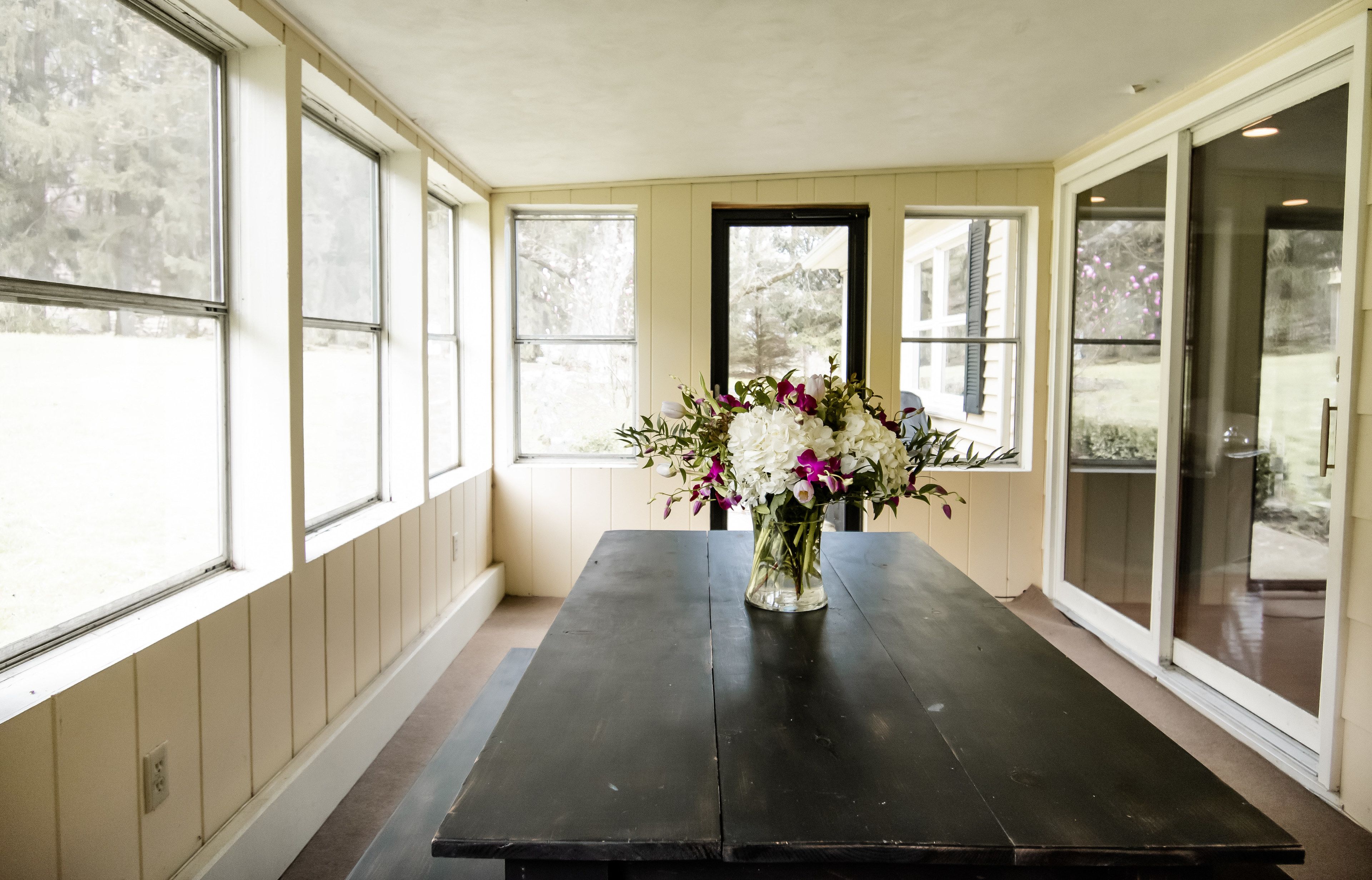 Um quarto com uma mesa de madeira escura, um vaso com flores e janelas grandes com vista para árvores.