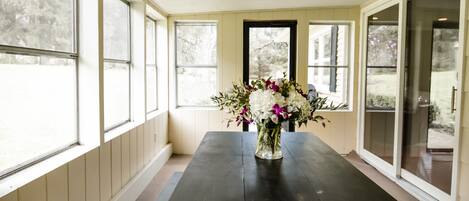 Una habitación con una mesa de madera oscura, un jarrón con flores y ventanas amplias que dan a un paisaje de árboles.