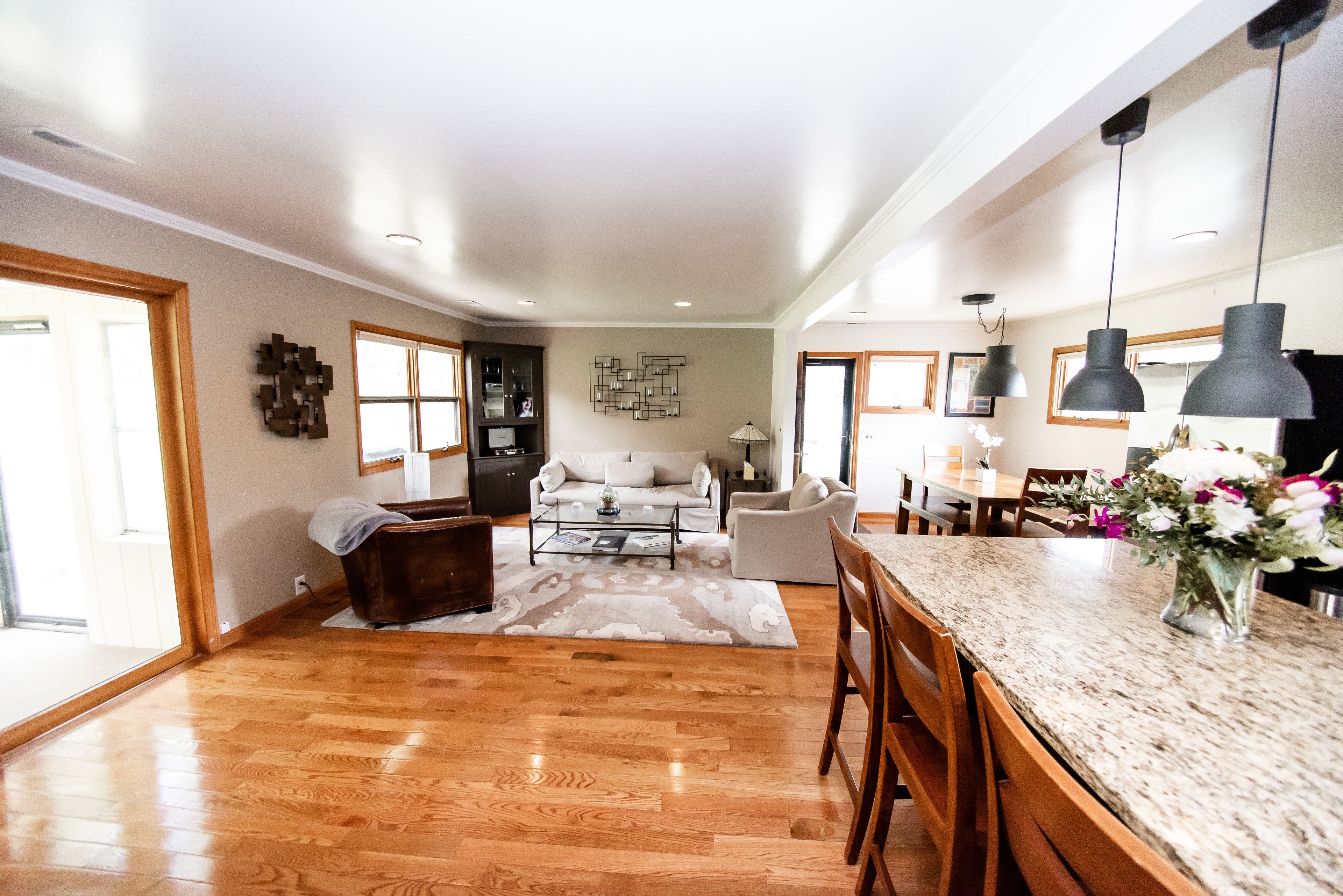 A spacious living area with a wooden floor, a white sofa, a coffee table, and a dining area with a marble countertop and bar stools.