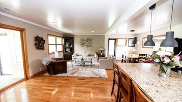 A spacious living area with a wooden floor, a white sofa, a coffee table, and a dining area with a marble countertop and bar stools.