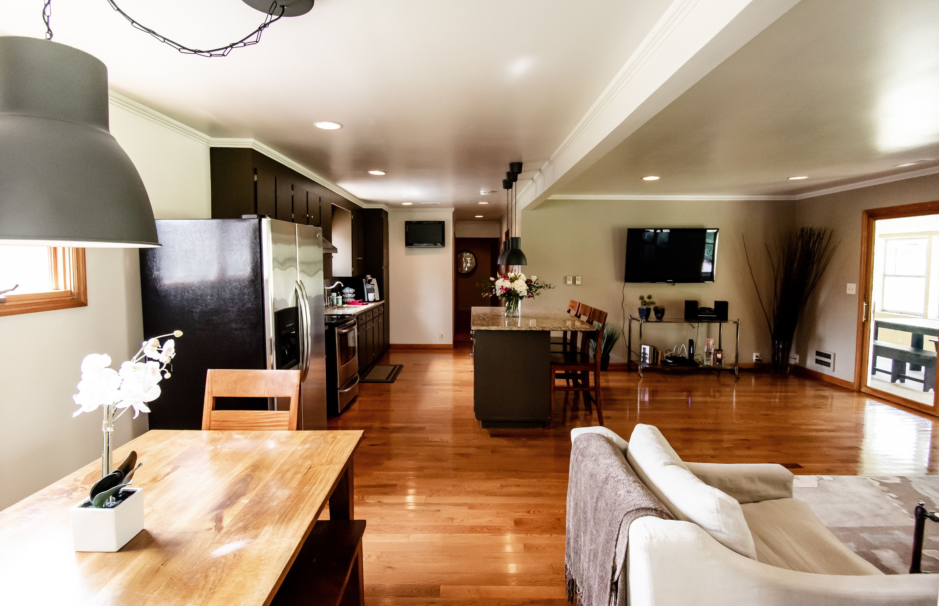 Un amplio salón con piso de madera, una mesa de comedor con sillas, un sofá, una cocina con armarios oscuros, un refrigerador y un televisor de pantalla plana montado en la pared.