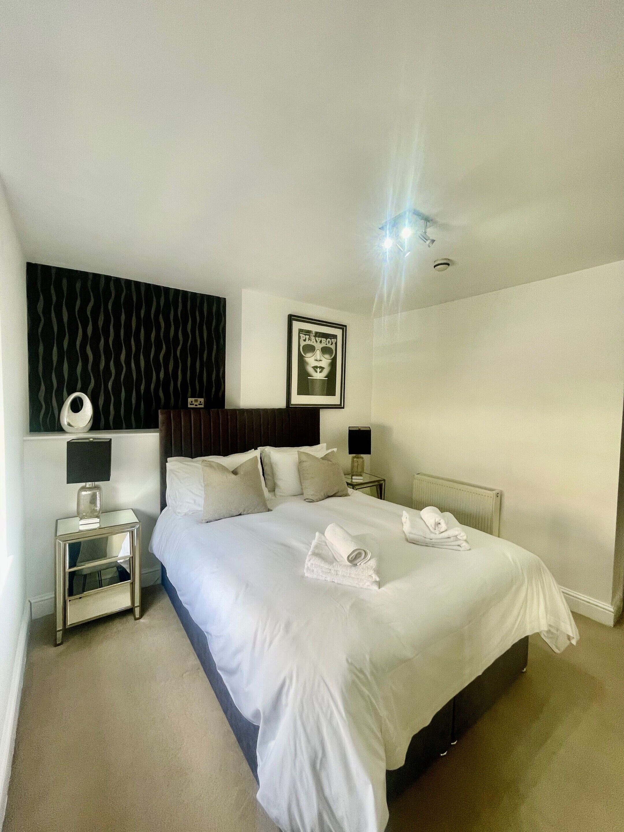 A hotel room with a neatly made bed, a bedside table with a lamp, a framed picture on the wall, and a radiator under the bed.