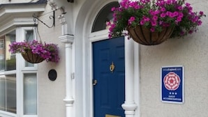 Une porte bleue avec une poignée dorée, une enseigne indiquant qu’il s’agit d’un logement pour les clients, et des jardinières suspendues.