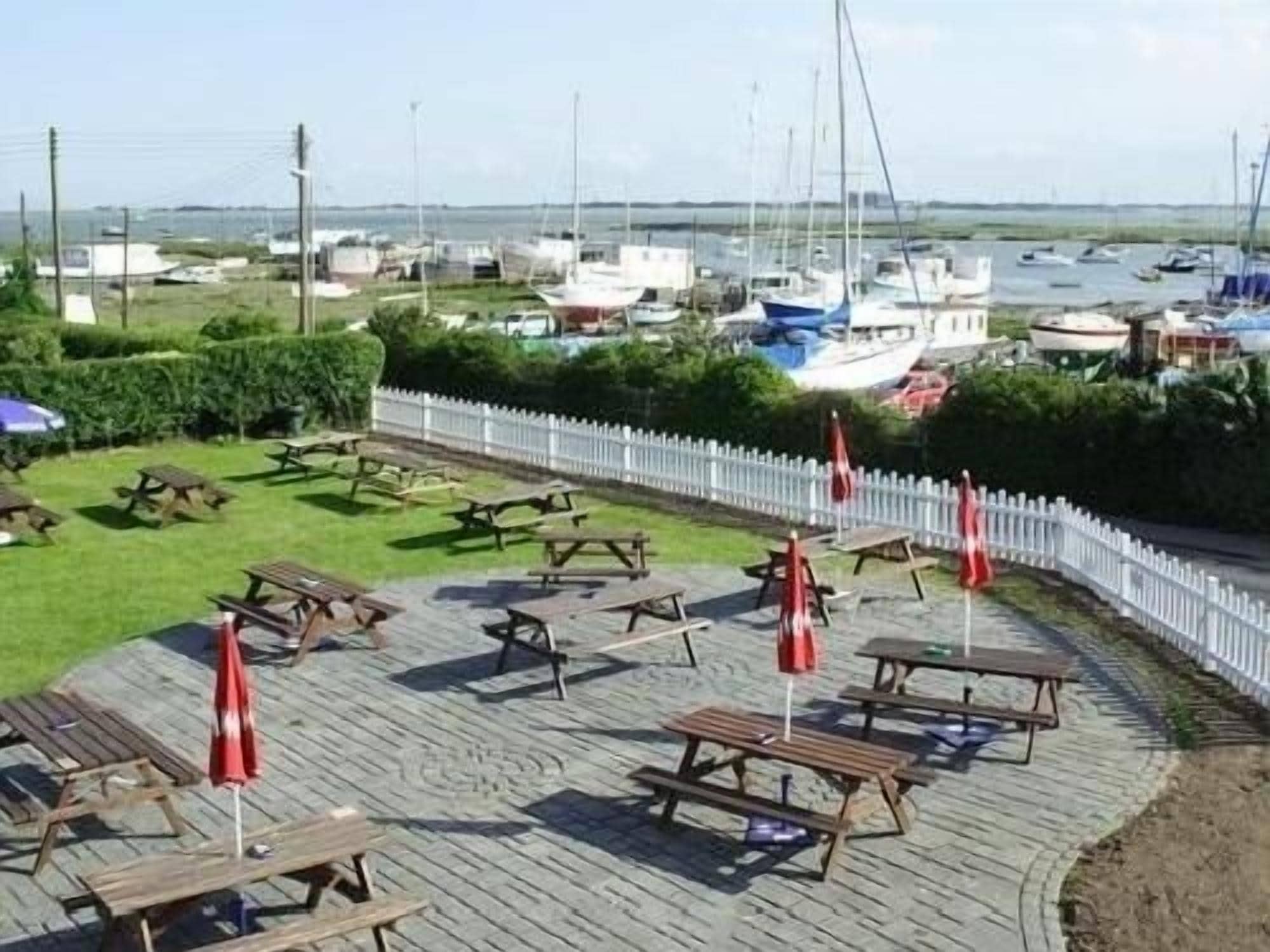 Un patio frente al agua con mesas de picnic y sombrillas, con vista a un puerto deportivo y barcos.