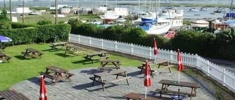 Un patio frente al agua con mesas de picnic y sombrillas, con vista a un puerto deportivo y barcos.