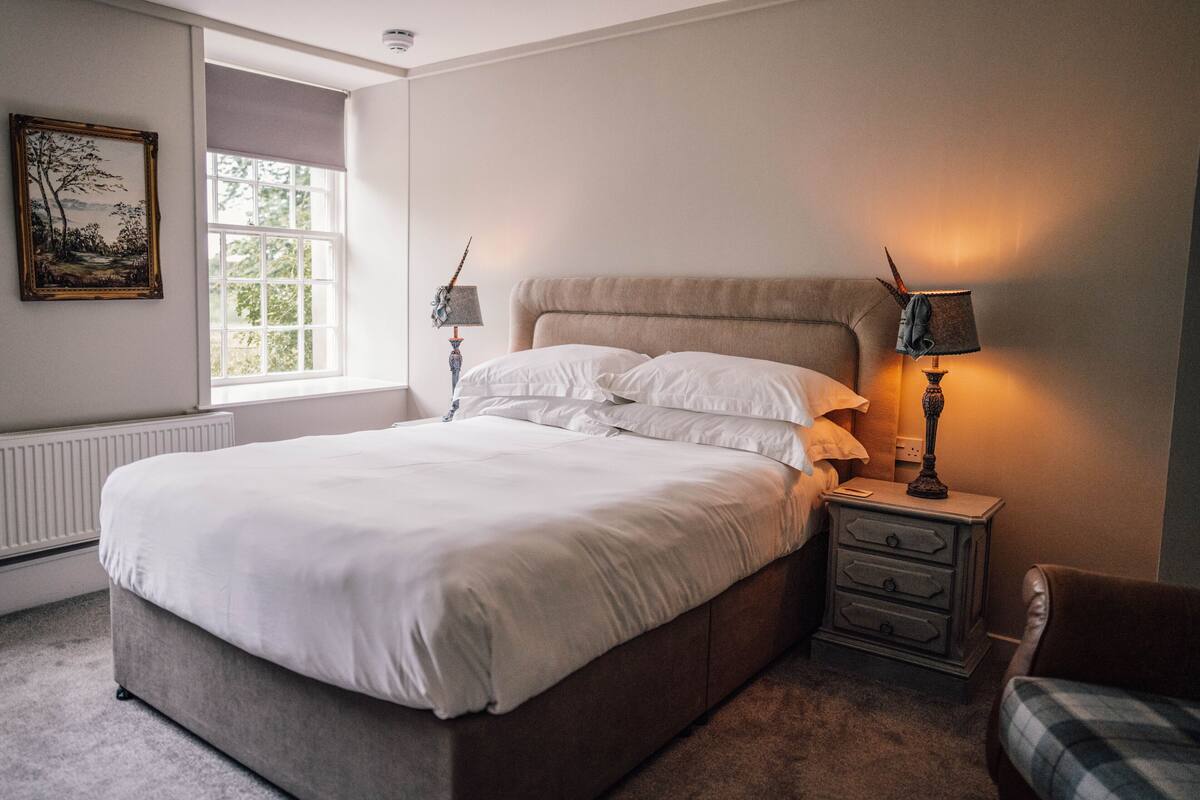 A bedroom with a large bed, two bedside tables, a chair, a window, and a painting on the wall.