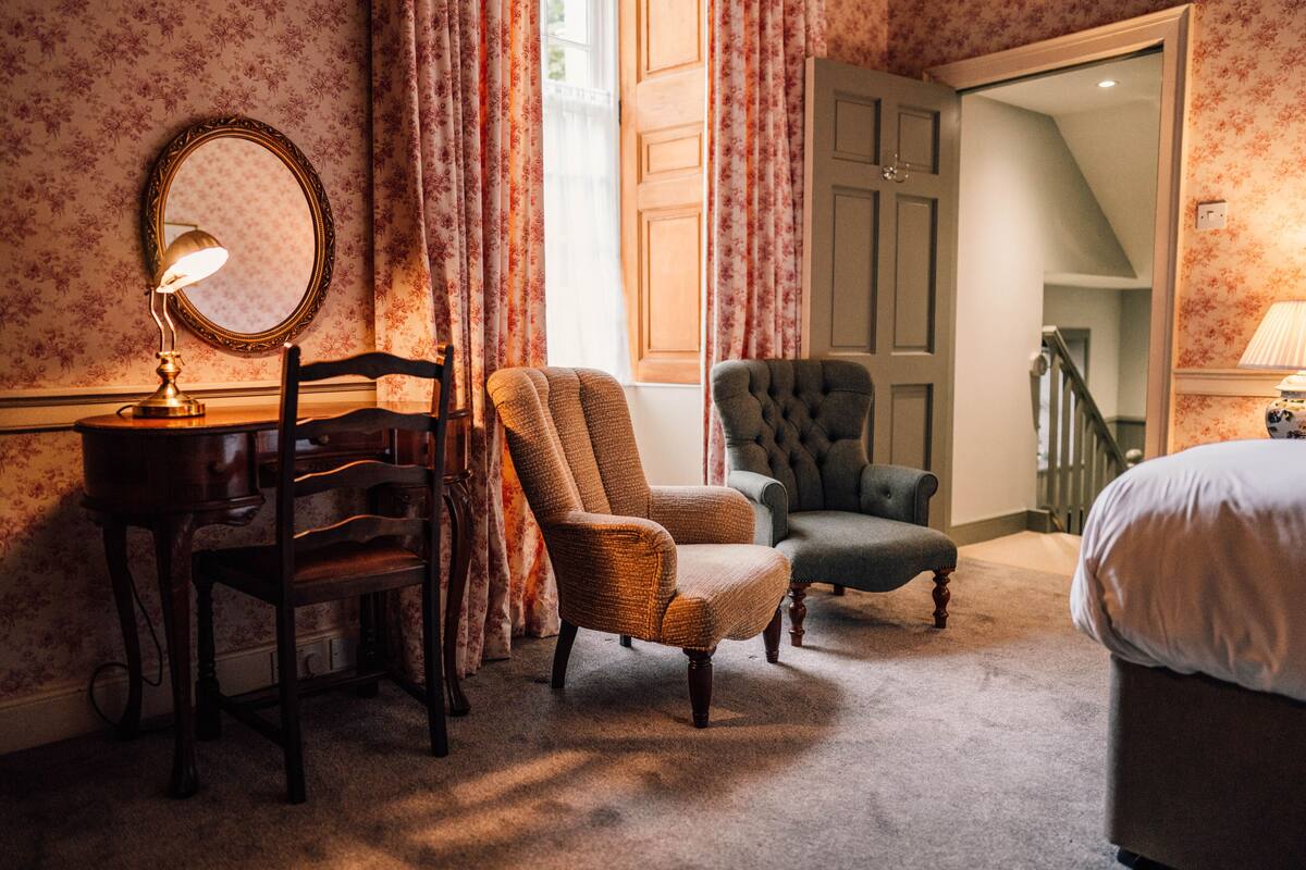 A bedroom with a bed, two armchairs, a wooden chair, a desk with a lamp, and a mirror.