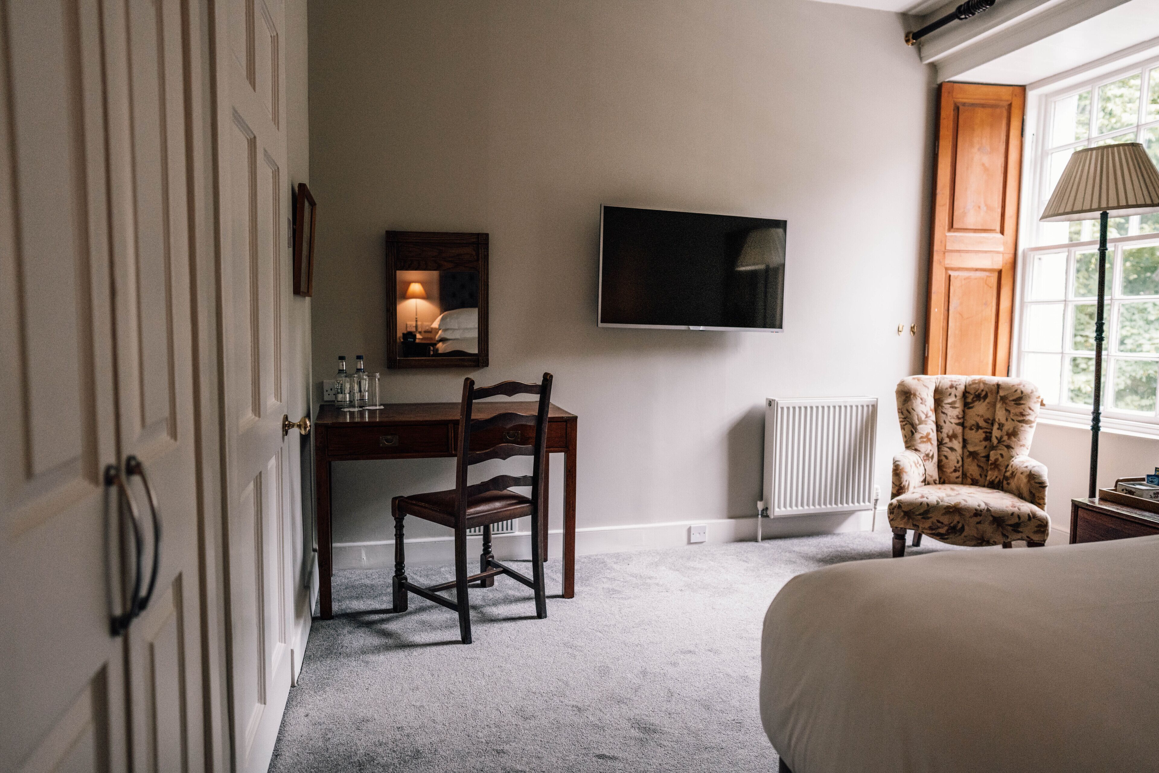 A bedroom with a bed, a desk with a chair, a television, a radiator, and a window with a lamp.