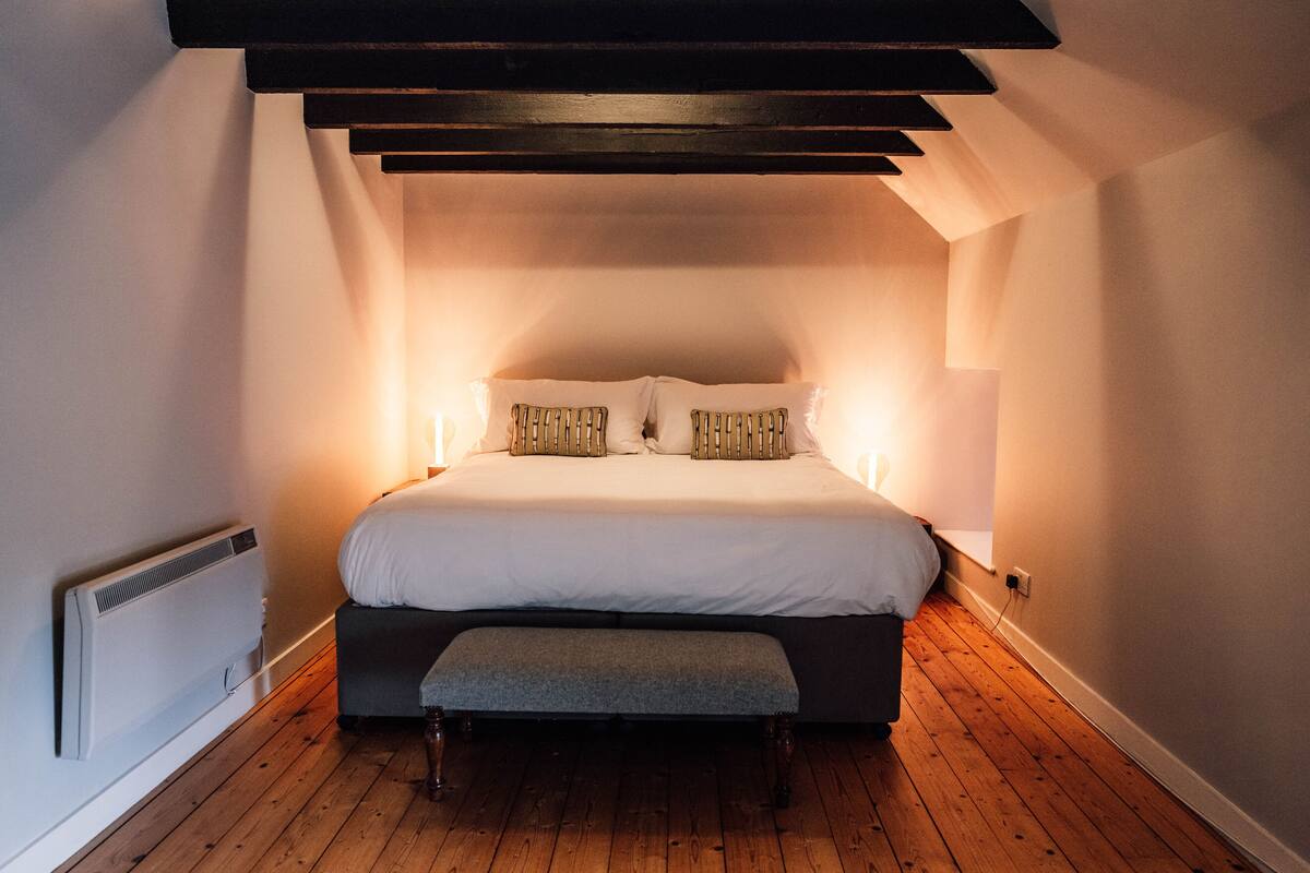 A bedroom with a wooden floor, a bed with white bedding, a bench at the foot of the bed, and a sloped ceiling with exposed beams.